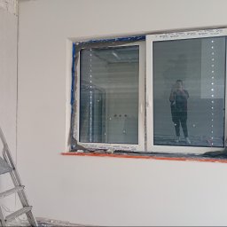 Agnieszka Jarosz Mama Na Sterydach - Ściana po szpachlowaniu z oknem zabezpieczonym folią, drabina oparta o ścianę. Widoczne ślady prac wykończeniowych. W oknie odbija się osoba robiąca zdjęcie.