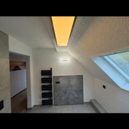 Agnieszka Jarosz Mama Na Sterydach - Wykończona łazienka na poddaszu z oknem dachowym, szarą płytką na ścianie i podłodze, grzejnikiem drabinkowym i oświetleniem sufitowym. Widok na sąsiednie pomieszczenie.