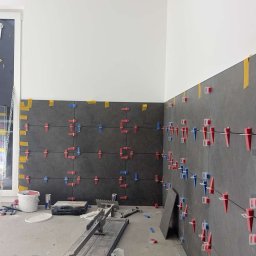 Agnieszka Jarosz Mama Na Sterydach - Układanie ciemnych płytek na ścianie z użyciem klipsów poziomujących. Widoczne narzędzia glazurnicze i fragment okna. Praca w toku, surowy stan.