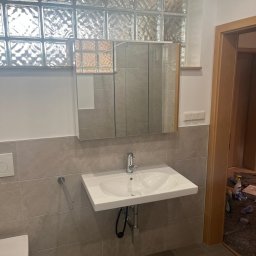 Mama Na Sterydach - Wykończona łazienka z umywalką, lustrem w szafce i luksferami nad nimi. Szare płytki na ścianach i podłodze. Widoczne elementy instalacji wodnej.