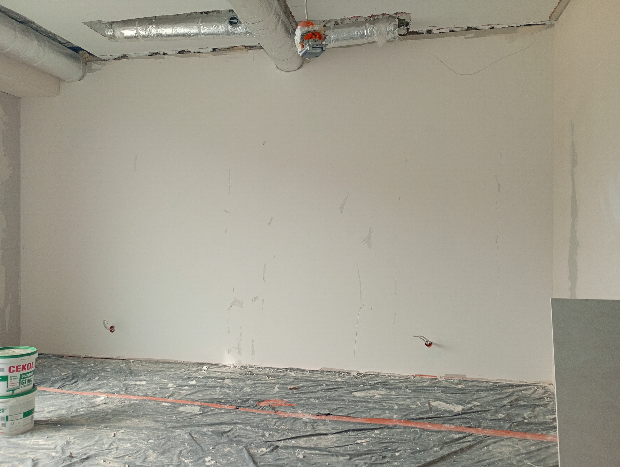 Biała ściana w trakcie szpachlowania, widoczne ślady po łatach, zabezpieczona podłoga folią, puszki elektryczne. Na pierwszym planie wiadro z masą szpachlową.