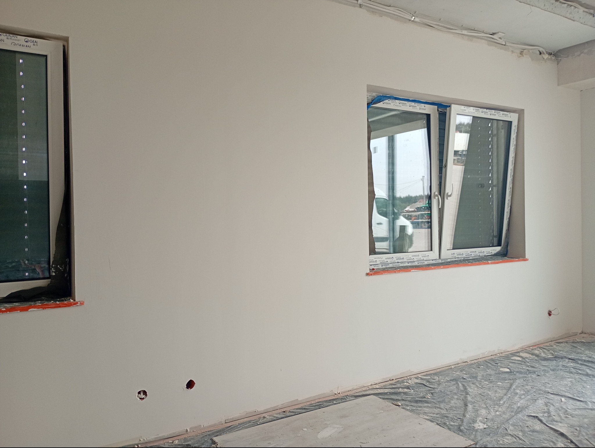 Biała ściana z dwoma oknami, jedno otwarte, drugie zasłonięte folią. Widoczne ślady po gniazdkach elektrycznych. Podłoga zabezpieczona folią malarską.