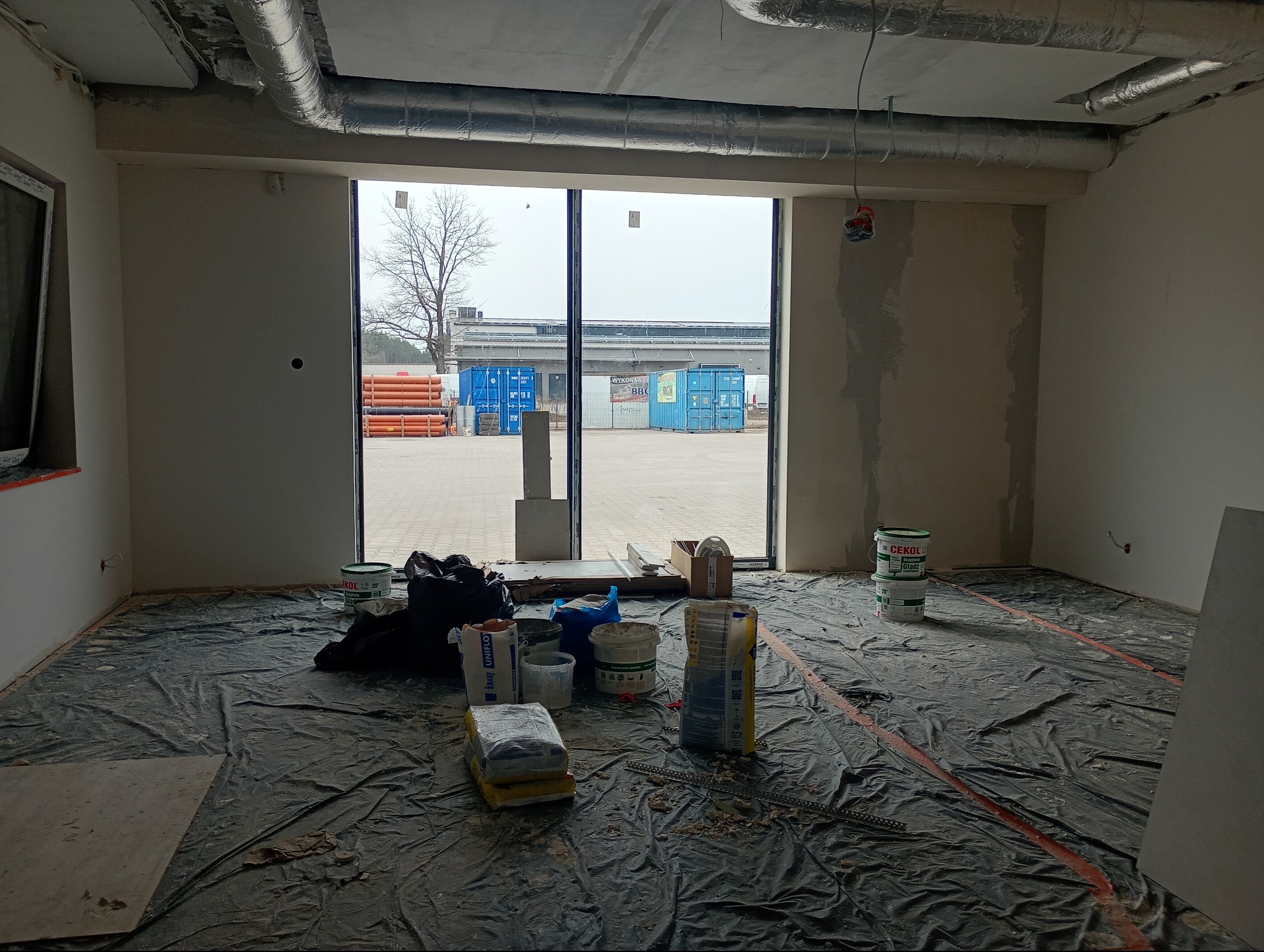 Wnętrze w trakcie prac przygotowawczych: podłoga zabezpieczona folią, widoczne materiały budowlane i częściowo wyszpachlowane ściany. Na zewnątrz widoczne kontenery.