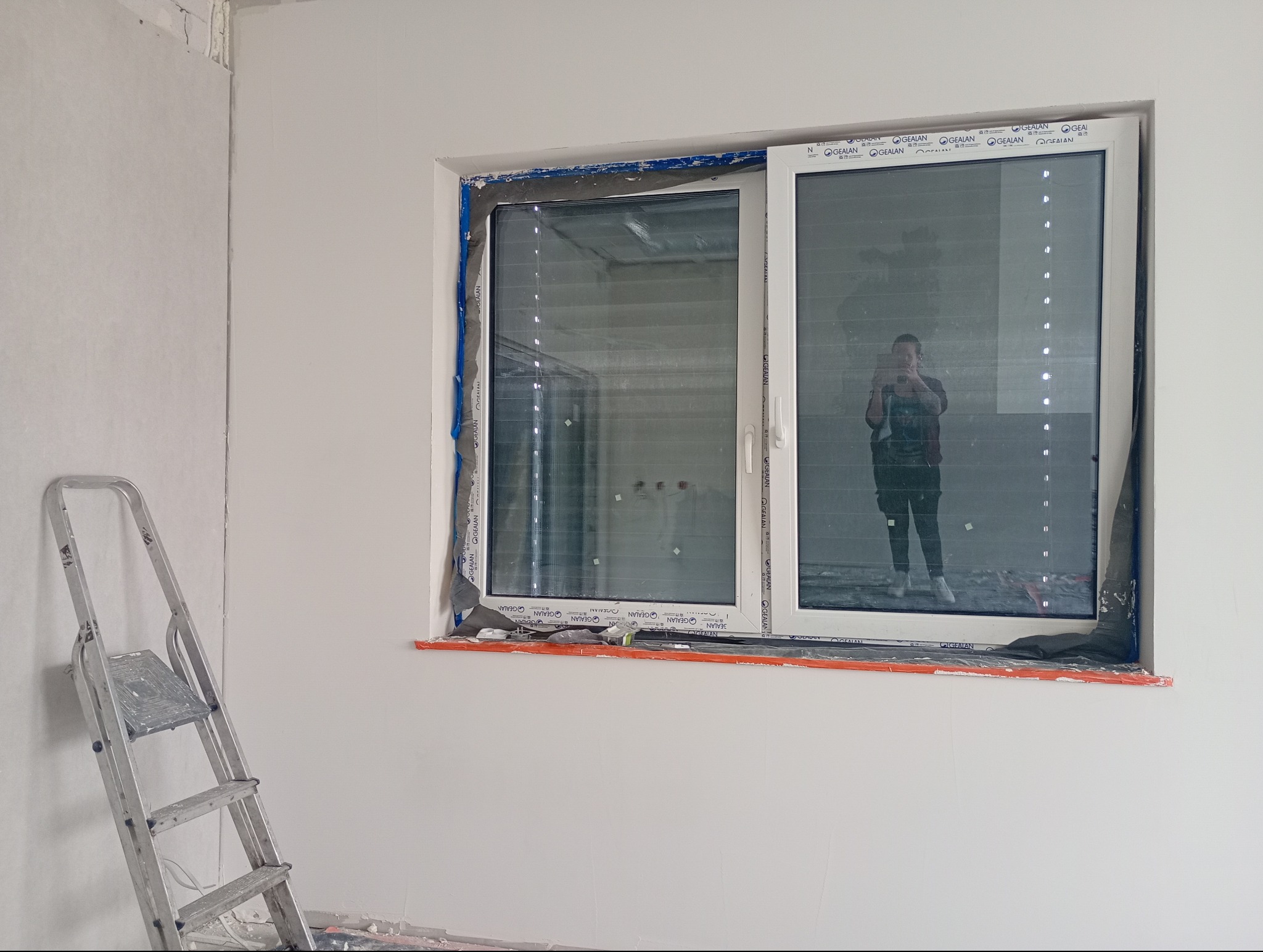 Ściana po szpachlowaniu z oknem zabezpieczonym folią, drabina oparta o ścianę. Widoczne ślady prac wykończeniowych. W oknie odbija się osoba robiąca zdjęcie.