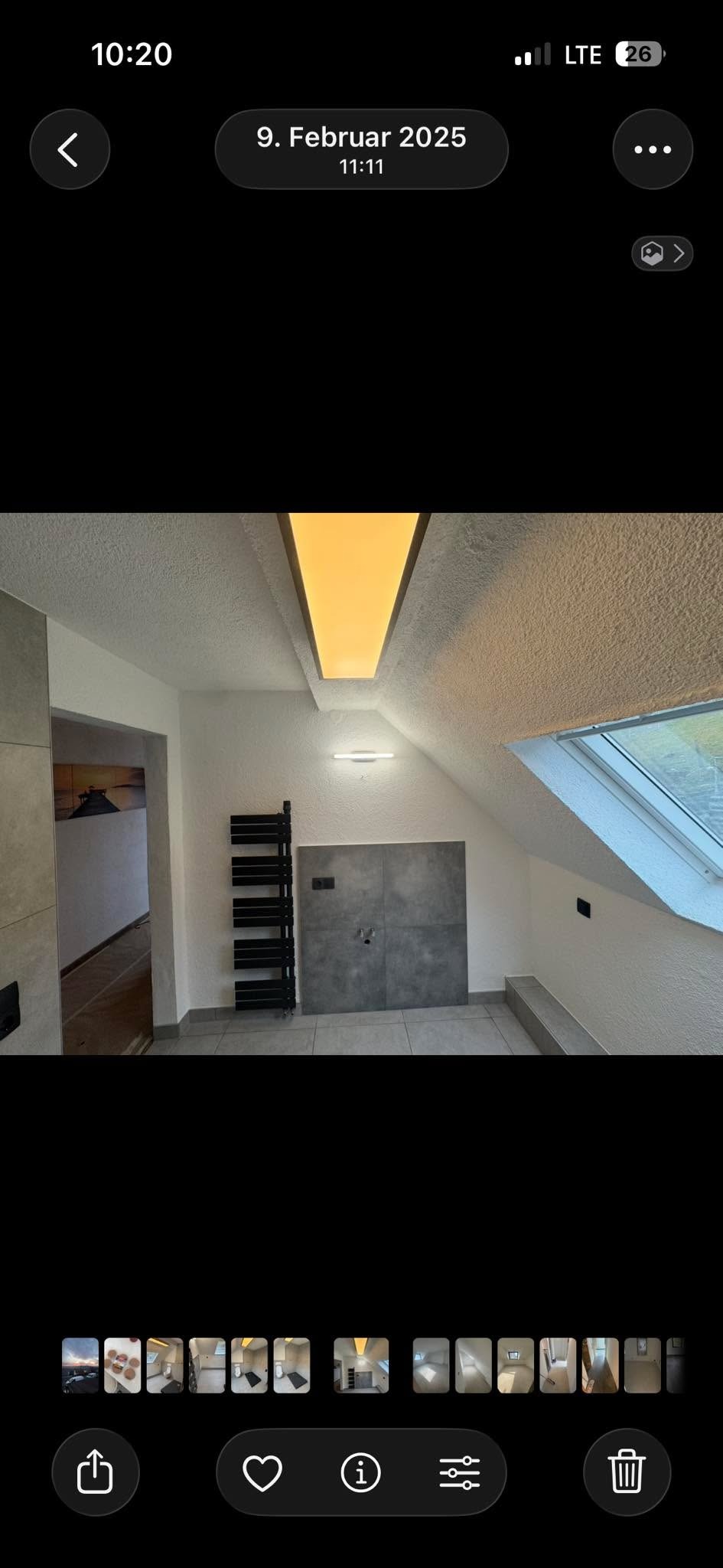 Wykończona łazienka na poddaszu z oknem dachowym, szarą płytką na ścianie i podłodze, grzejnikiem drabinkowym i oświetleniem sufitowym. Widok na sąsiednie pomieszczenie.