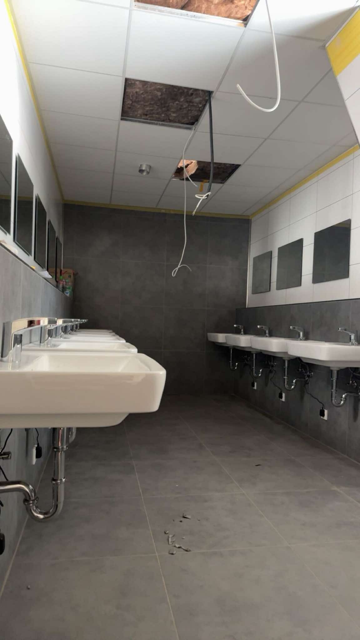 Wnętrze łazienki w trakcie remontu: rzędy umywalek, szare płytki, częściowo zdjęty sufit podwieszany z widoczną izolacją. Widoczne kable i elementy instalacji.