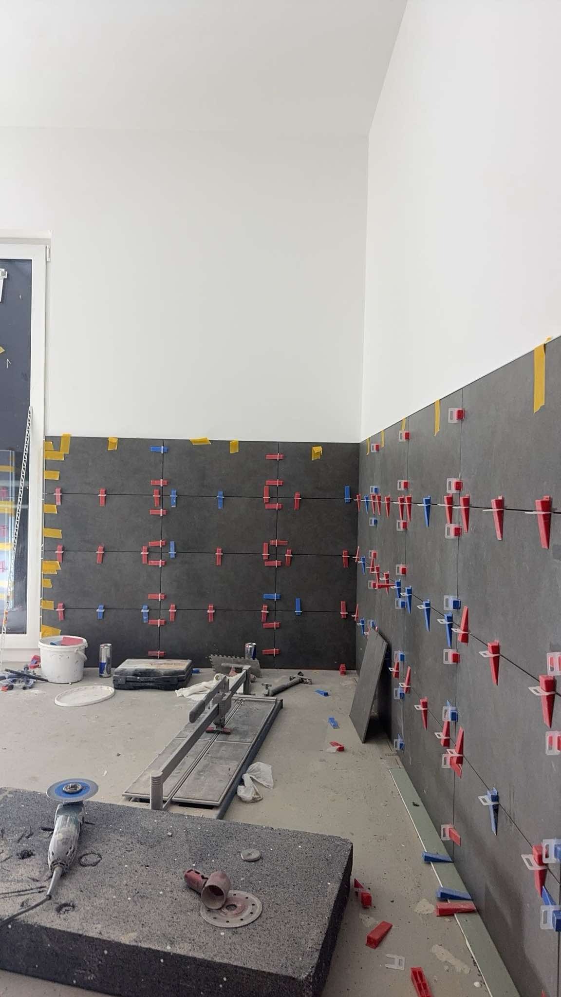 Układanie ciemnych płytek na ścianie z użyciem klipsów poziomujących. Widoczne narzędzia glazurnicze i fragment okna. Praca w toku, surowy stan.