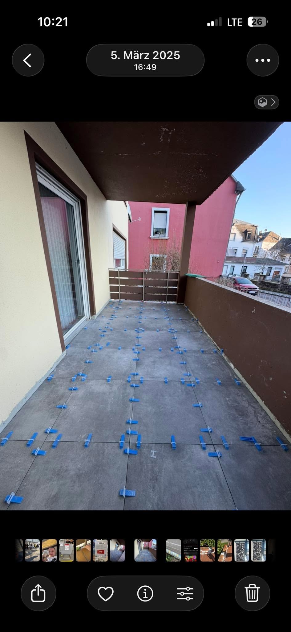 Szare płytki na balkonie w trakcie układania, z użyciem niebieskich krzyżyków dystansowych. Widok na budynki w tle.