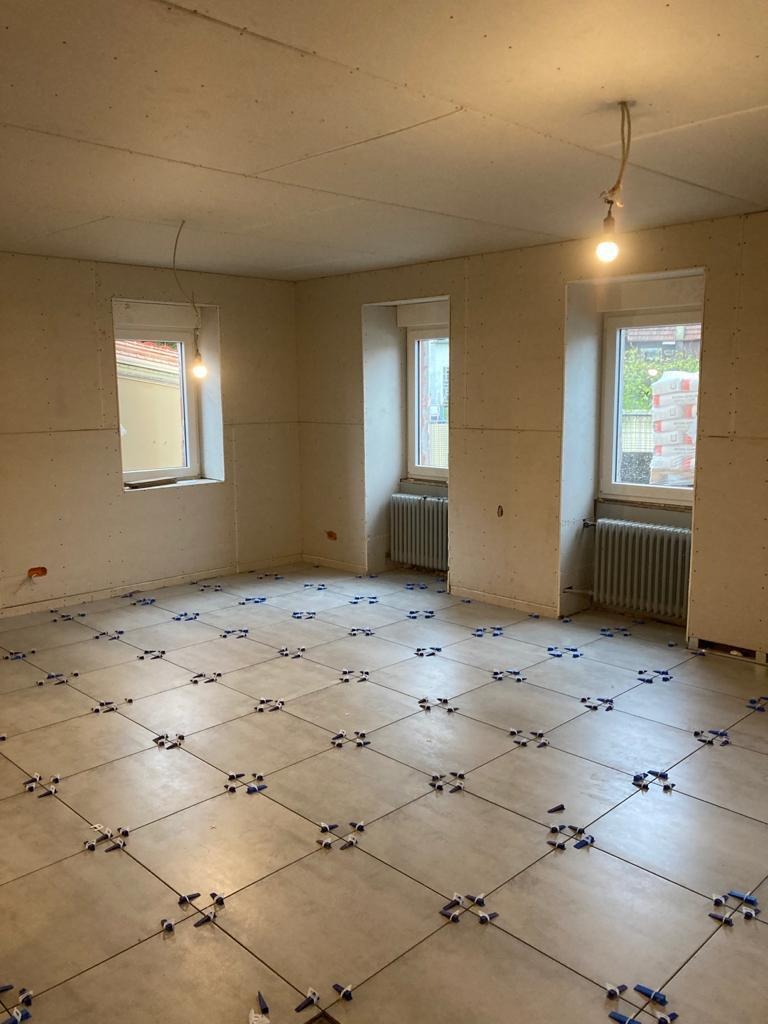 Wnętrze w trakcie układania płytek podłogowych z użyciem krzyżyków dystansowych. Widoczne okna z grzejnikami i surowy sufit z żarówką.