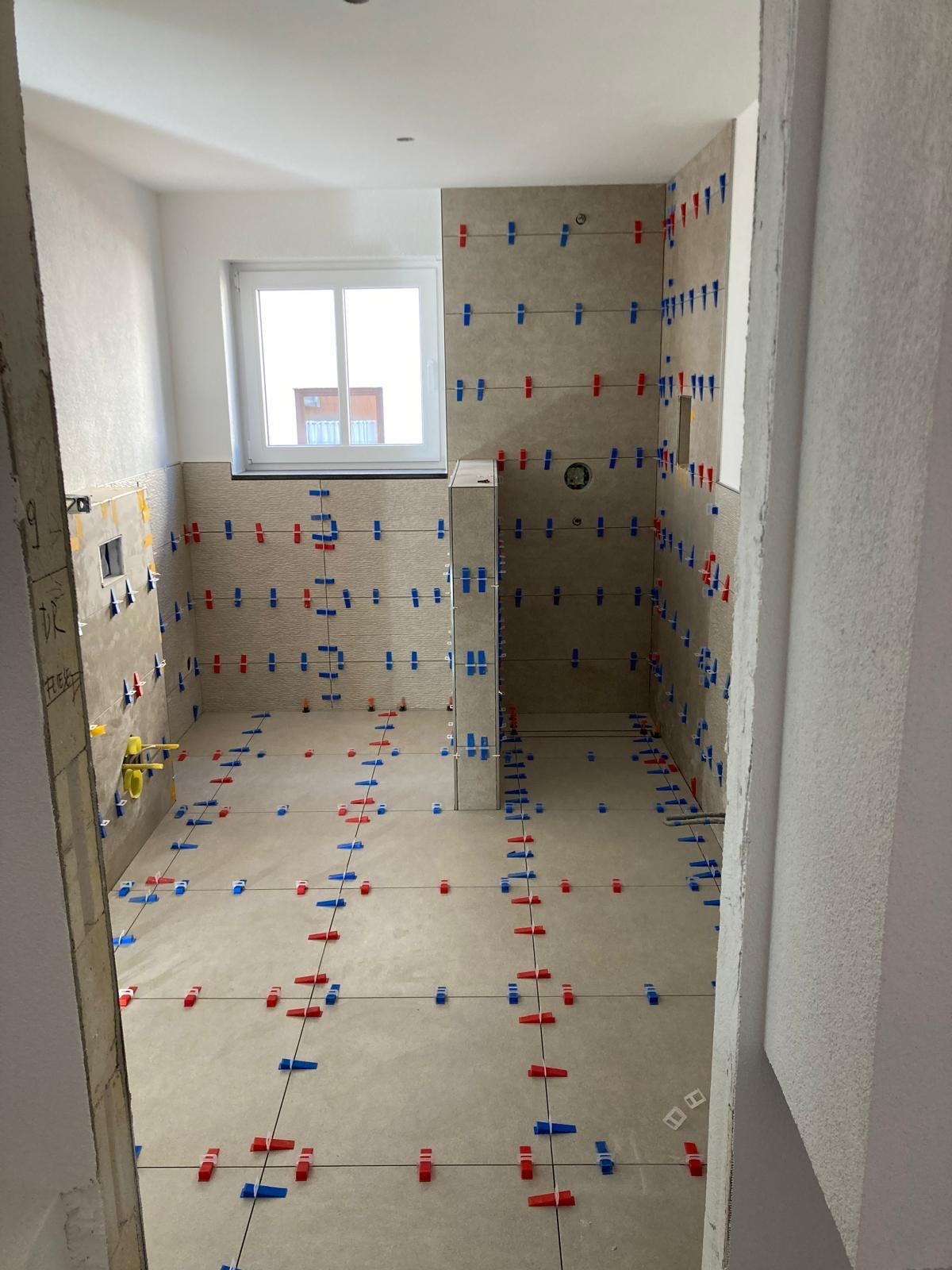 Układanie płytek w łazience z użyciem klipsów poziomujących. Widoczne świeżo położone płytki na ścianach i podłodze, przygotowane do fugowania. Jasne wnętrze z oknem.