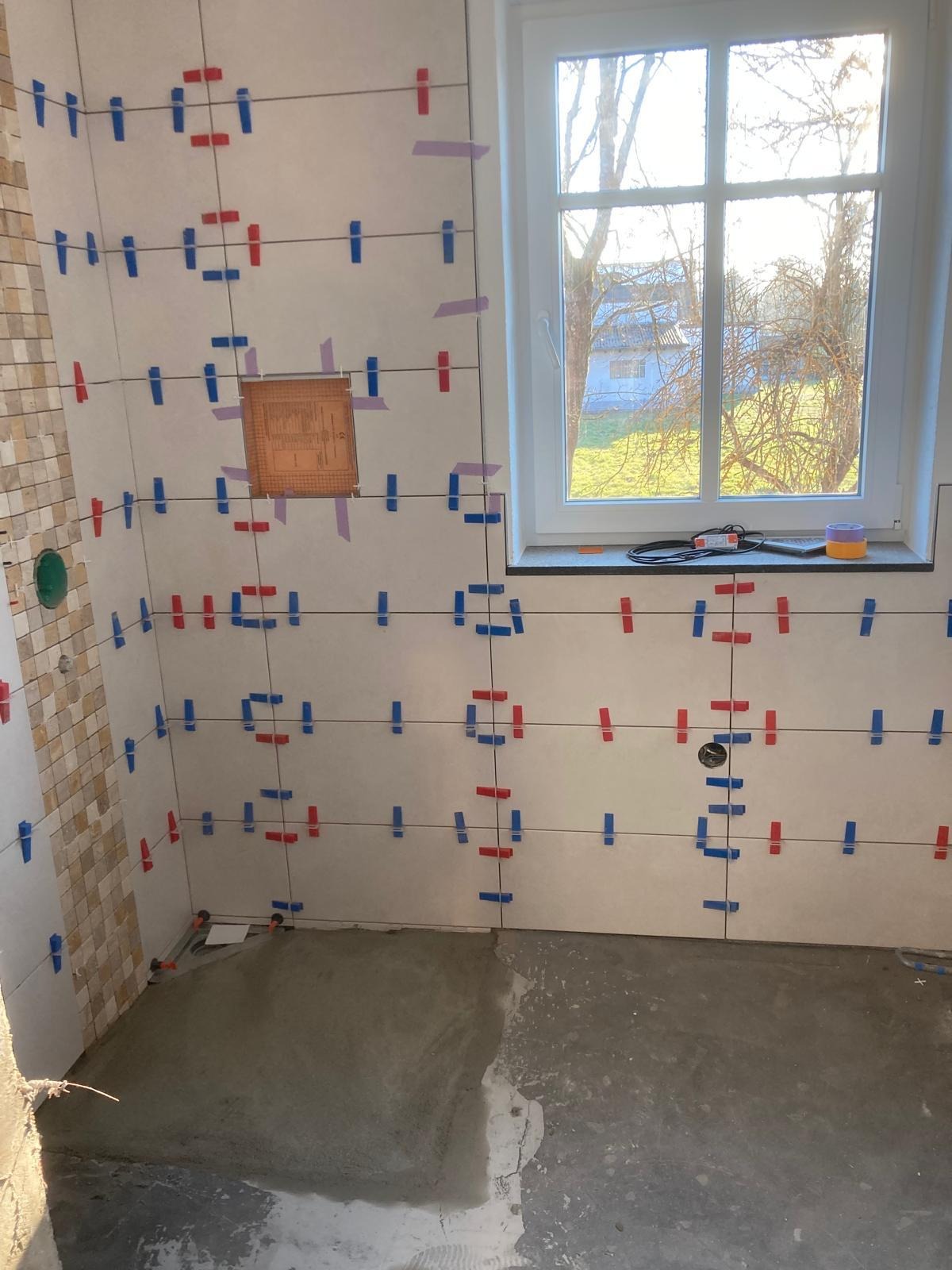 Układanie płytek w łazience, widoczne płytki z niebieskimi i czerwonymi znacznikami, okno z widokiem na zewnątrz, częściowo ułożona mozaika na ścianie.