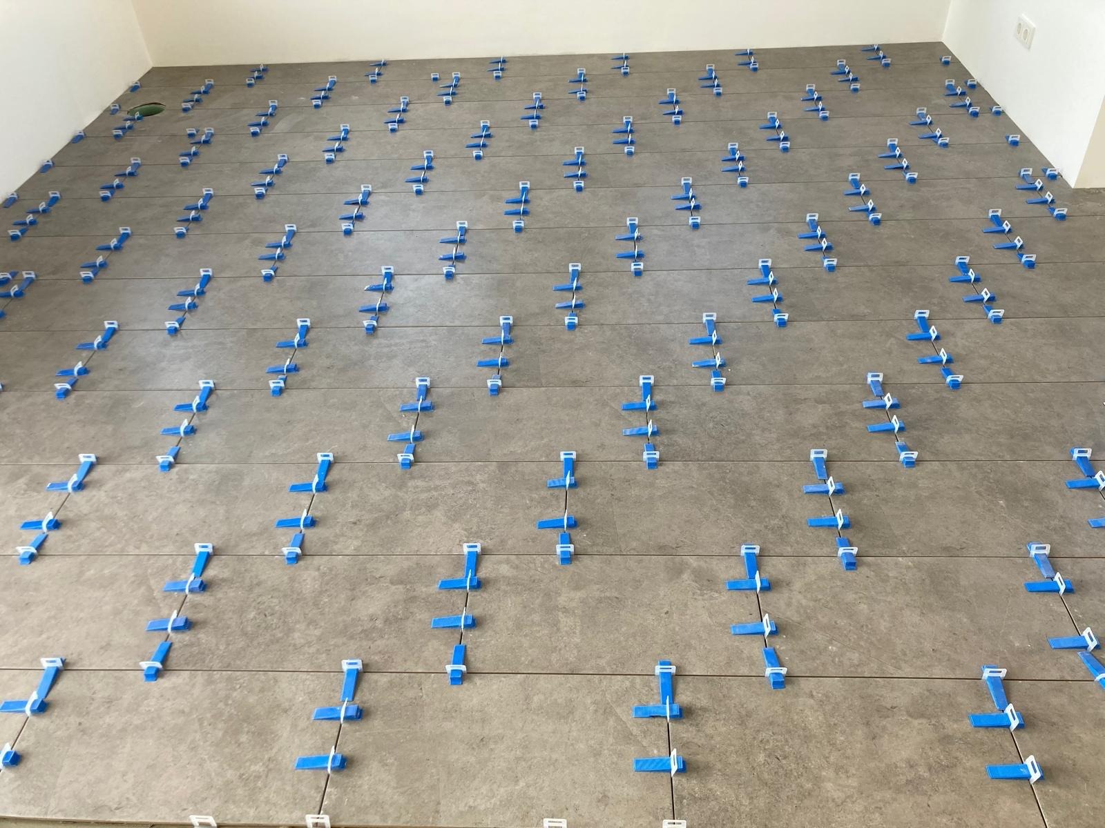 Ułożone, szare płytki podłogowe z niebieskimi klipsami poziomującymi, widok z góry. Etap prac przed fugowaniem, w jasnym pomieszczeniu.