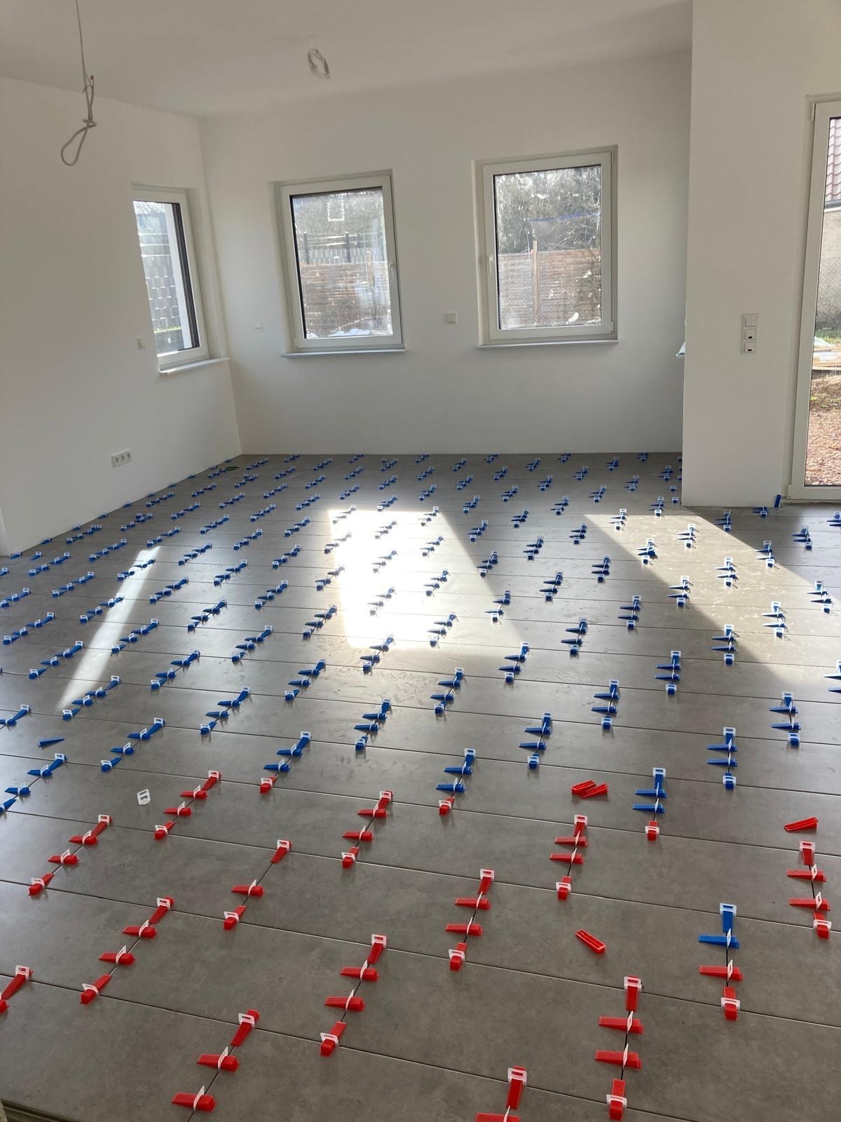 Układanie szarych płytek podłogowych z użyciem niebieskich i czerwonych klipsów poziomujących w jasnym, nowym pomieszczeniu z trzema oknami.