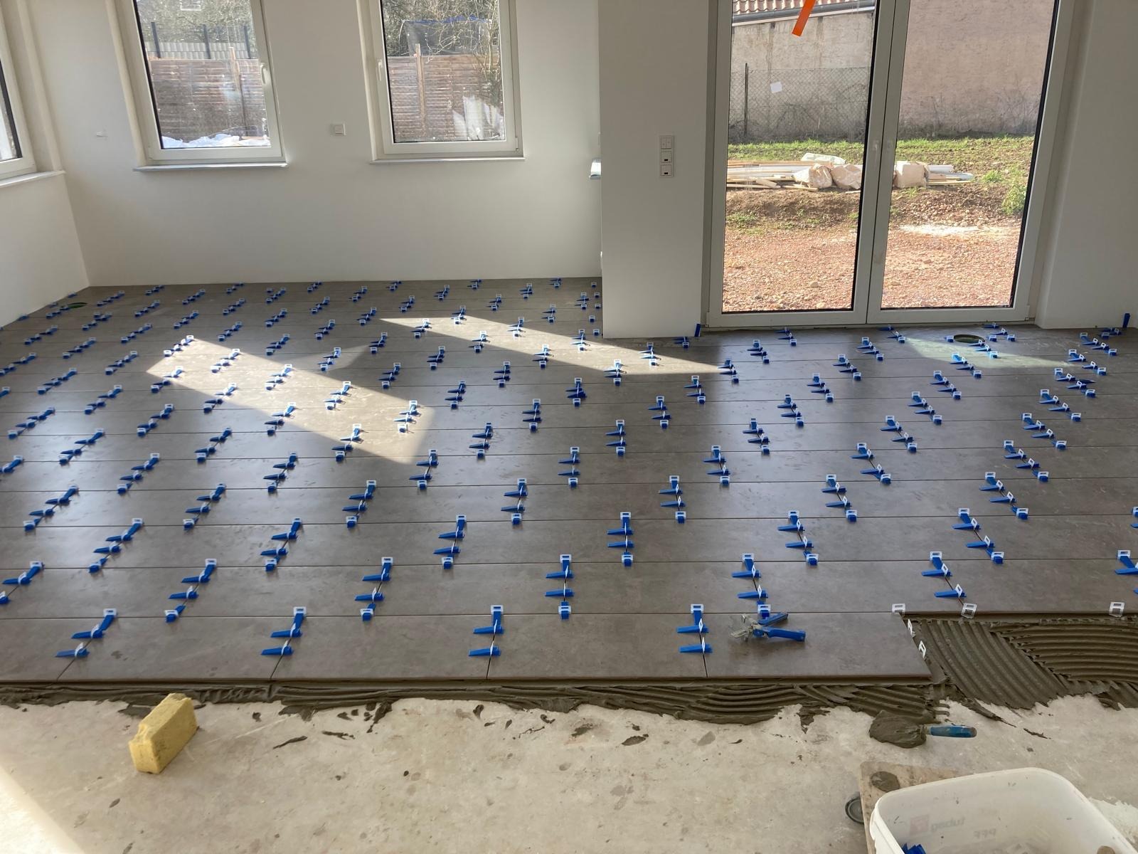 Układanie szarych płytek podłogowych z użyciem niebieskich klipsów poziomujących w jasnym pomieszczeniu z oknami i drzwiami balkonowymi.