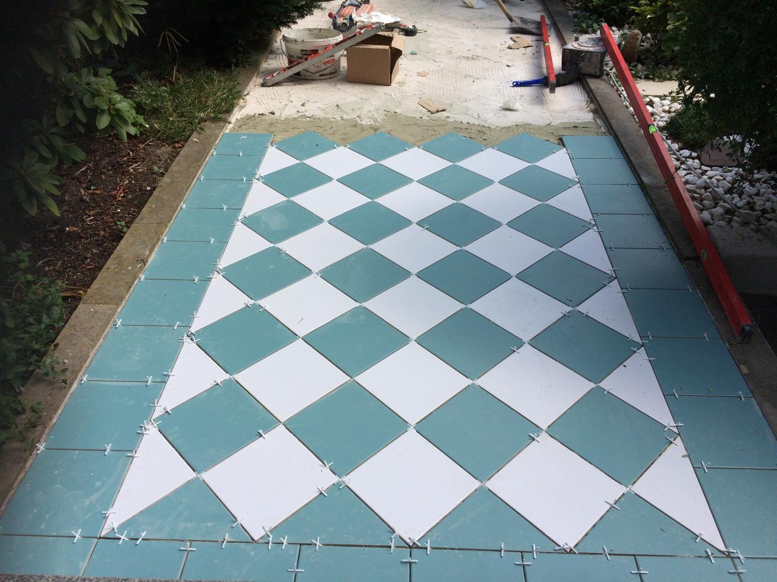 Układanie wzoru w romby z białych i turkusowych płytek na zewnątrz, widoczne krzyżyki dystansowe i narzędzia glazurnicze w tle.