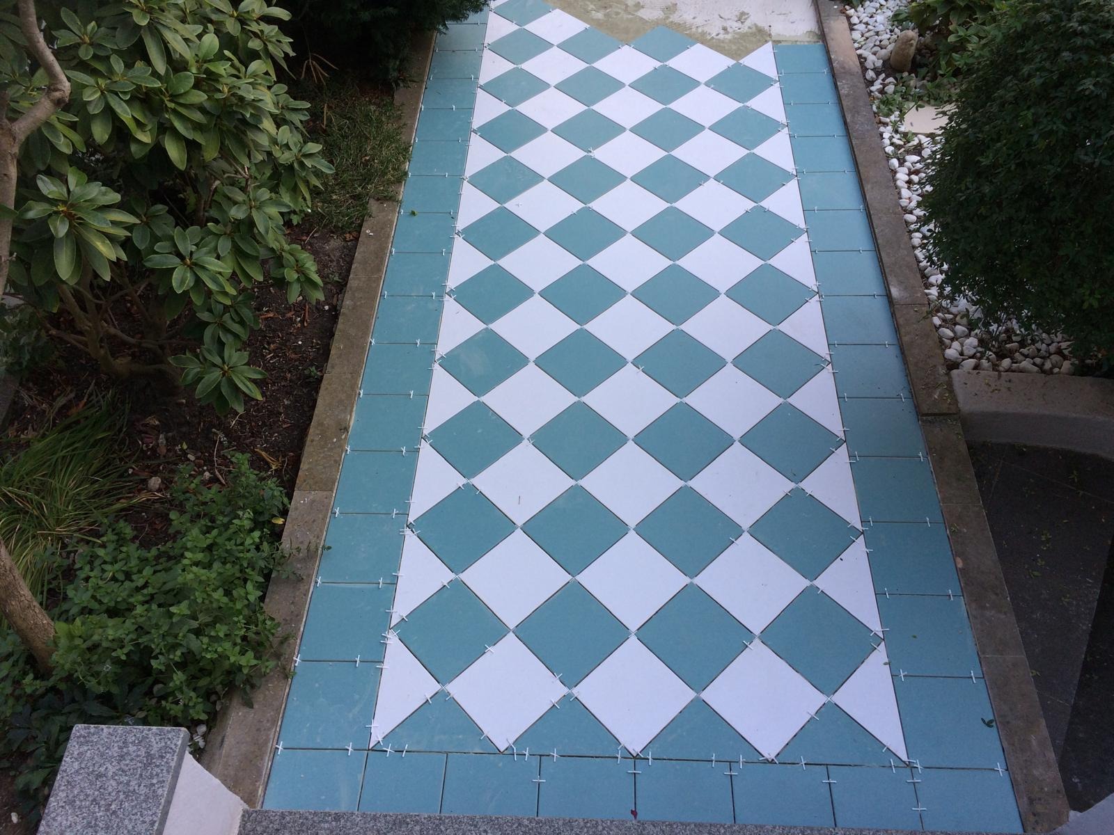 Ułożona podłoga z kwadratowych, biało-turkusowych płytek w romby, z widocznymi krzyżykami dystansowymi, otoczona zielenią. Ujęcie z góry.