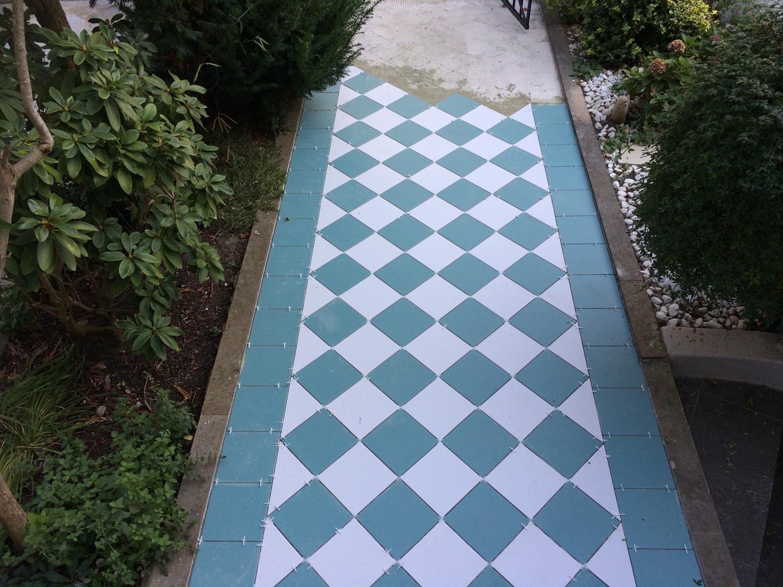 Ułożona, wzorzysta posadzka z białych i turkusowych płytek w romby, z krzyżykami dystansowymi, otoczona zielenią. Częściowo ukończona, z widocznym fragmentem starej nawierzchni.