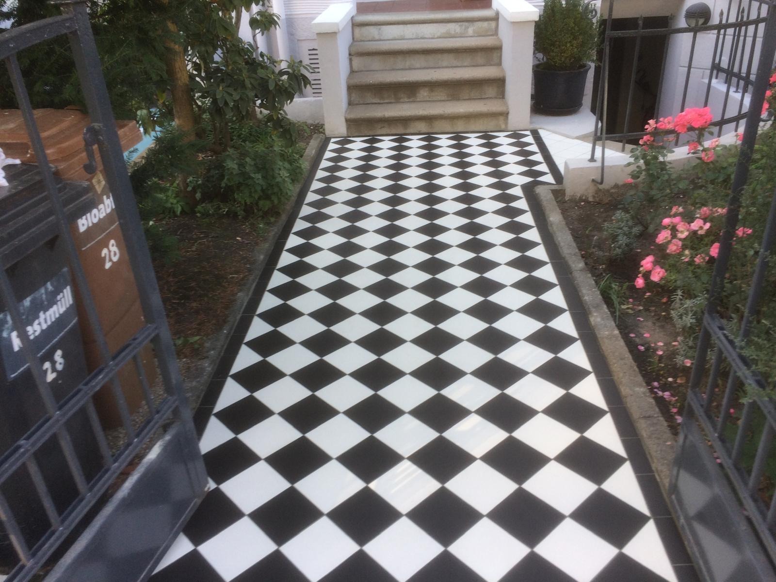 Ułożona czarno-biała szachownica z płytek prowadząca do schodów wejściowych, widok zza otwartej furtki, zieleń po bokach. Klasyczny wzór na zewnątrz budynku.
