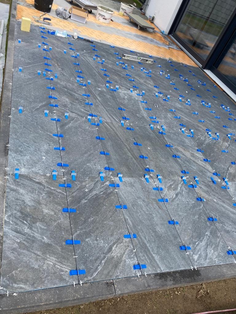 Układanie szarych płytek na tarasie z użyciem niebieskich klipsów poziomujących. Widoczne narzędzia i materiały budowlane. Prace glazurnicze w toku.