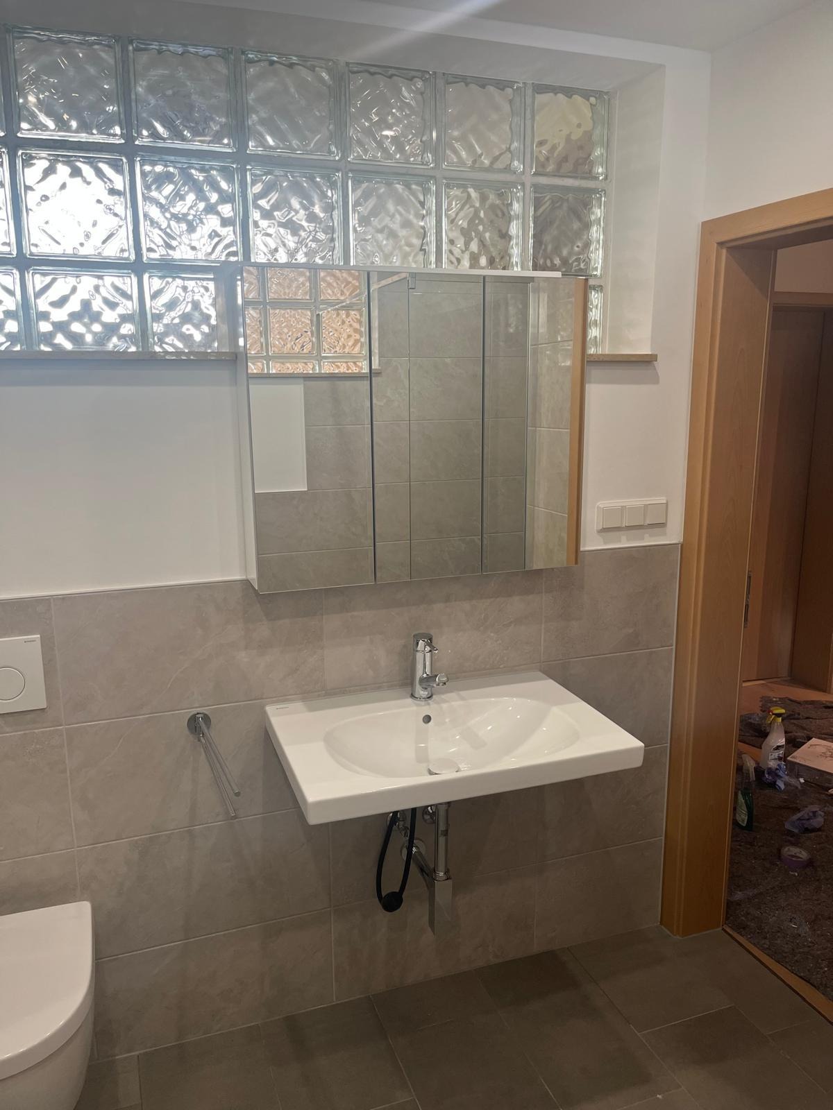 Wykończona łazienka z umywalką, lustrem w szafce i luksferami nad nimi. Szare płytki na ścianach i podłodze. Widoczne elementy instalacji wodnej.