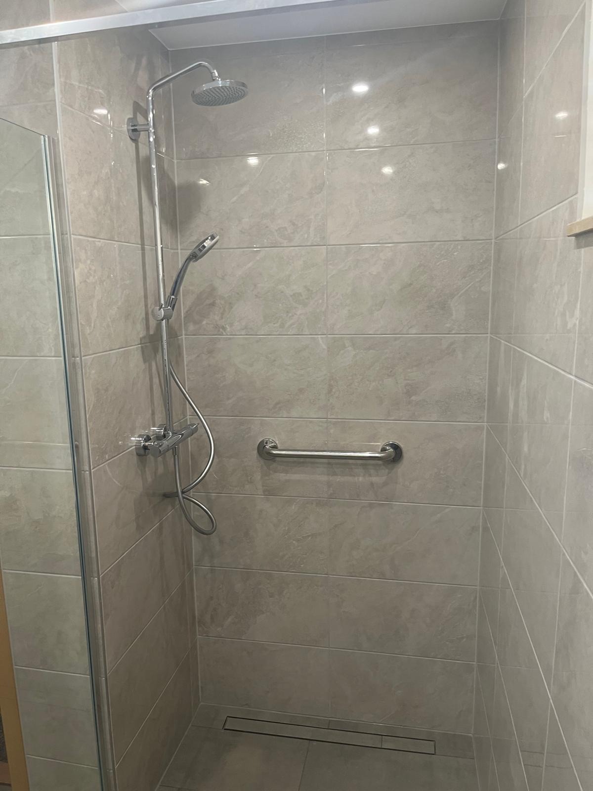 Wykończona kabina prysznicowa z szarymi płytkami imitującymi marmur, chromowaną armaturą i uchwytem dla niepełnosprawnych. Nowoczesny design z odpływem liniowym.