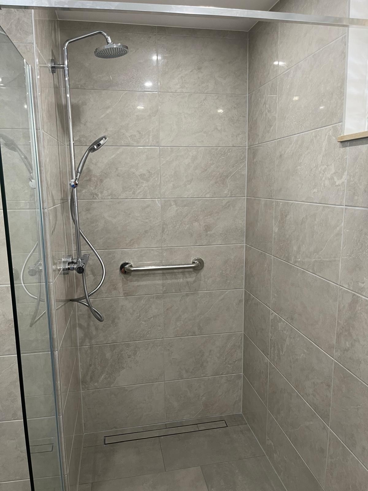 Nowoczesna kabina prysznicowa z szarymi płytkami imitującymi marmur, chromowaną armaturą i uchwytem dla niepełnosprawnych. Minimalistyczny design, odpływ liniowy.