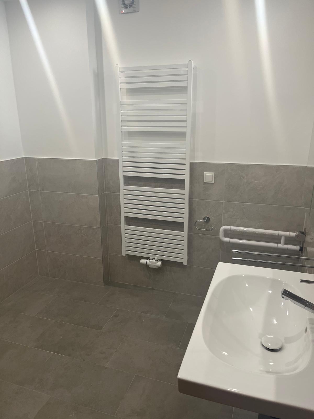 Wykończona łazienka w szarościach: płytki imitujące kamień, biała umywalka i grzejnik drabinkowy. Nowoczesny design z elementami dla osób z niepełnosprawnościami.