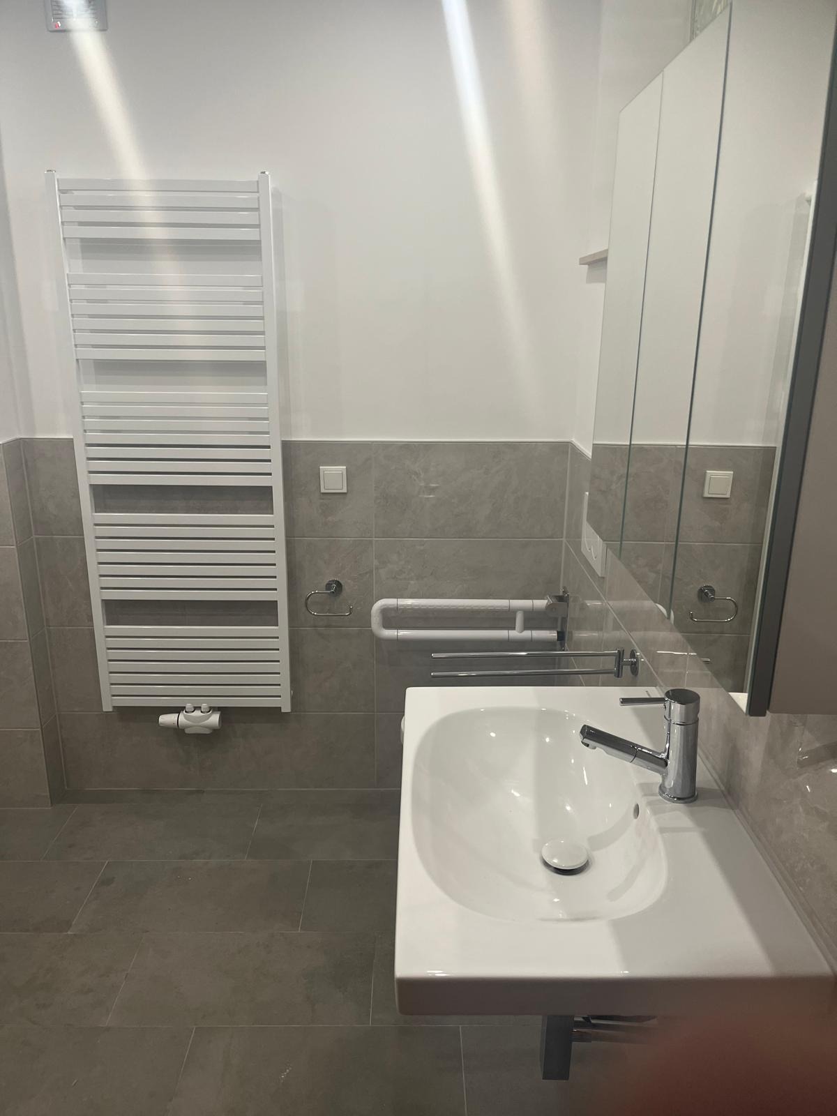 Wykończona łazienka w Białymstoku: biała umywalka, grzejnik drabinkowy i szare płytki. Nowoczesny design z uchwytami dla niepełnosprawnych.