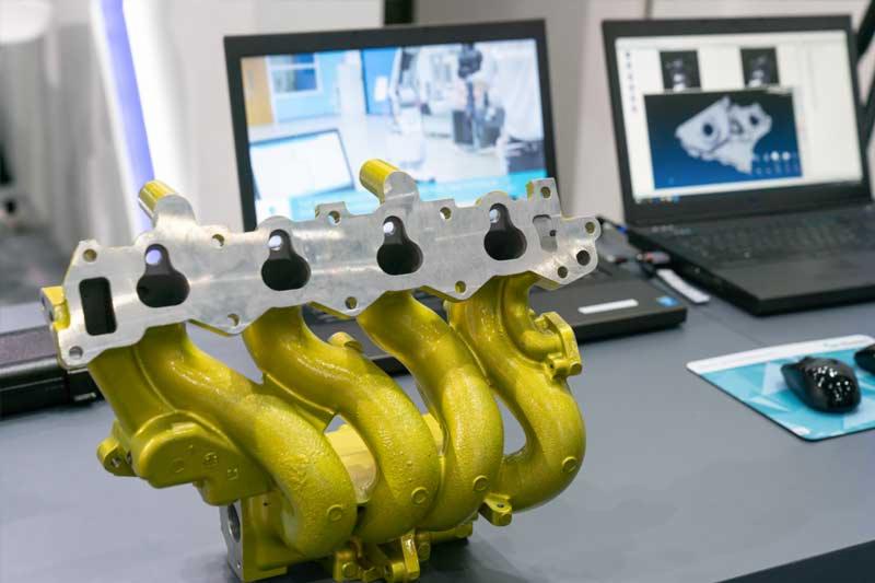 Żółty kolektor wydechowy z elementami obróbki skrawaniem, widoczne dwa laptopy z programami do projektowania CAD/CAM w tle.