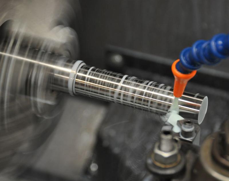 Metalowy wałek obrabiany w tokarce z użyciem chłodziwa, widoczne ślady obróbki w postaci regularnej siatki nacięć.