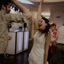 Panna młoda w koronie z kwiatów i białej sukni tańczy z uniesioną ręką, w tle DJ na podświetlonym stanowisku i inni goście weselni.