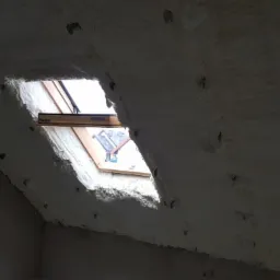 Poddasze z oknem dachowym, pokryte grubą warstwą białej pianki izolacyjnej z widocznymi otworami wentylacyjnymi, częściowo oświetlone światłem dziennym.