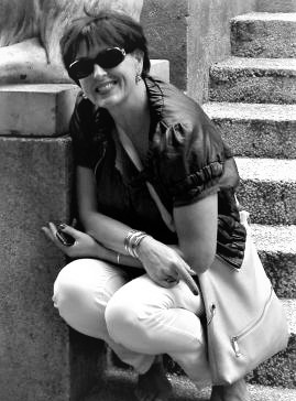 Uśmiechnięta kobieta w okularach przeciwsłonecznych kuca przy kamiennych schodach i rzeźbie lwa, wskazując palcem w dół. Zdjęcie czarno-białe.