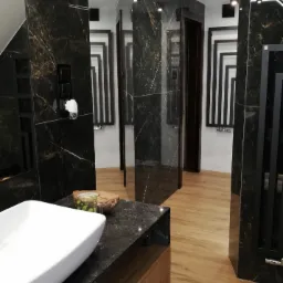 Elegancka łazienka z czarnymi marmurowymi płytkami, drewnianą podłogą i blatem, białą umywalką oraz dekoracyjnymi elementami w postaci czarnego grzejnika i szklanki z napojem na blacie.