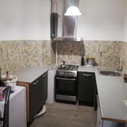 Wykończona kuchnia z patchworkowymi płytkami nad blatem, szarymi szafkami i sprzętami AGD, widok z wejścia do pomieszczenia.