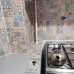 Kuchnia z patchworkowymi płytkami ceramicznymi na ścianie i nad blatem, srebrny czajnik na kuchence gazowej.