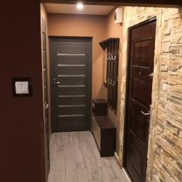 RB Bojarowski - Przedpokój z dwiema parami drzwi, wieszakiem z półką i dekoracyjną ścianą z kamienia, oświetlony górnym światłem.