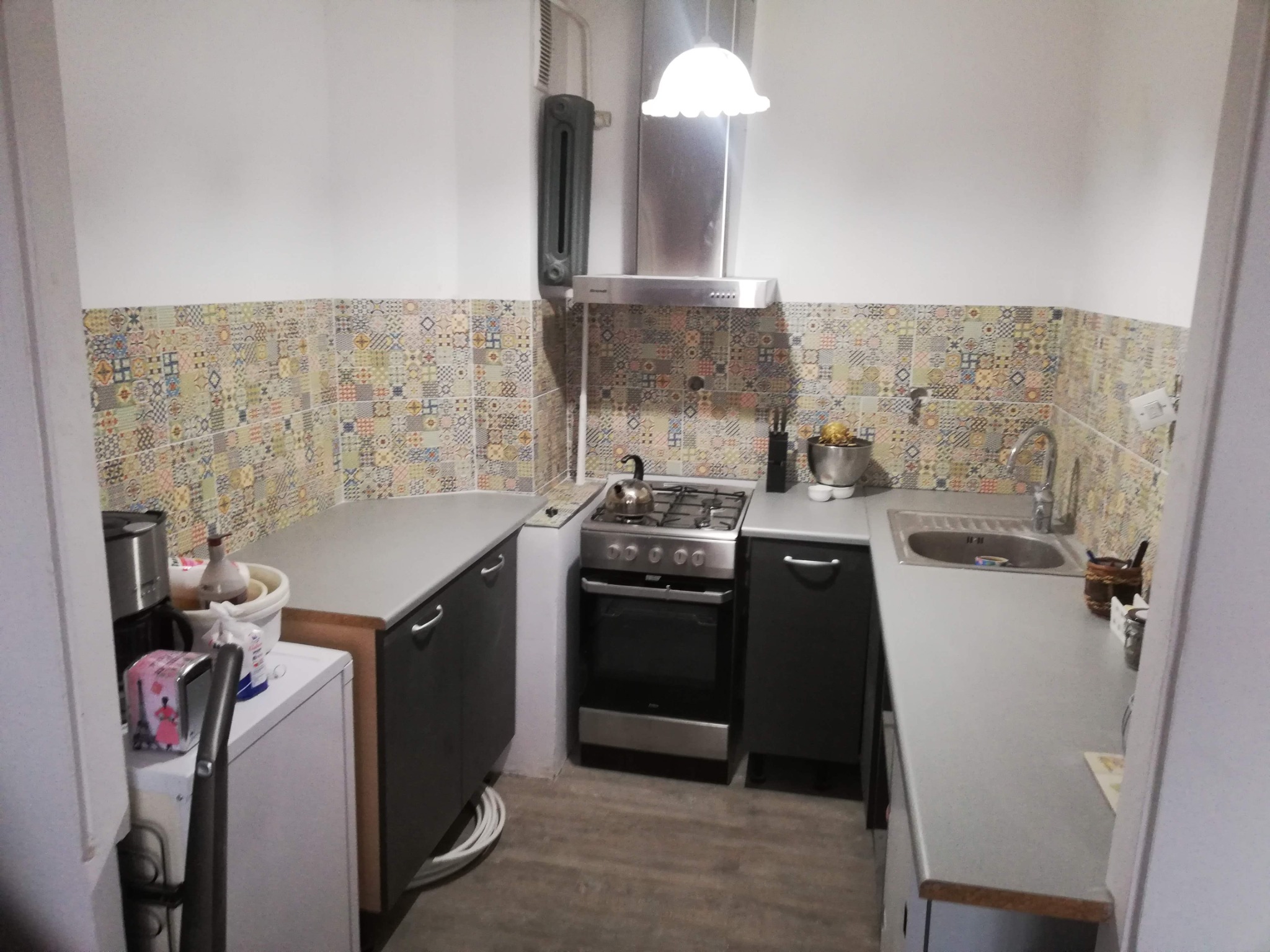 Wykończona kuchnia z patchworkowymi płytkami nad blatem, szarymi szafkami i sprzętami AGD, widok z wejścia do pomieszczenia.