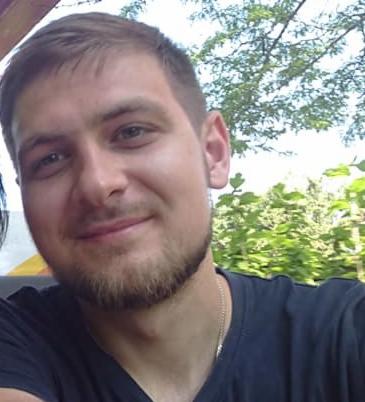 Uśmiechnięty mężczyzna z krótką brodą i srebrnym łańcuszkiem na tle zieleni i fragmentu nieba. Portret w naturalnym świetle.