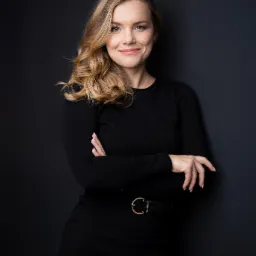 Portret kobiety z blond włosami, ubranej w czarną sukienkę z paskiem z okrągłą klamrą, stojącej na tle ciemnoszarej ściany, z założonymi rękami i subtelnym uśmiechem.