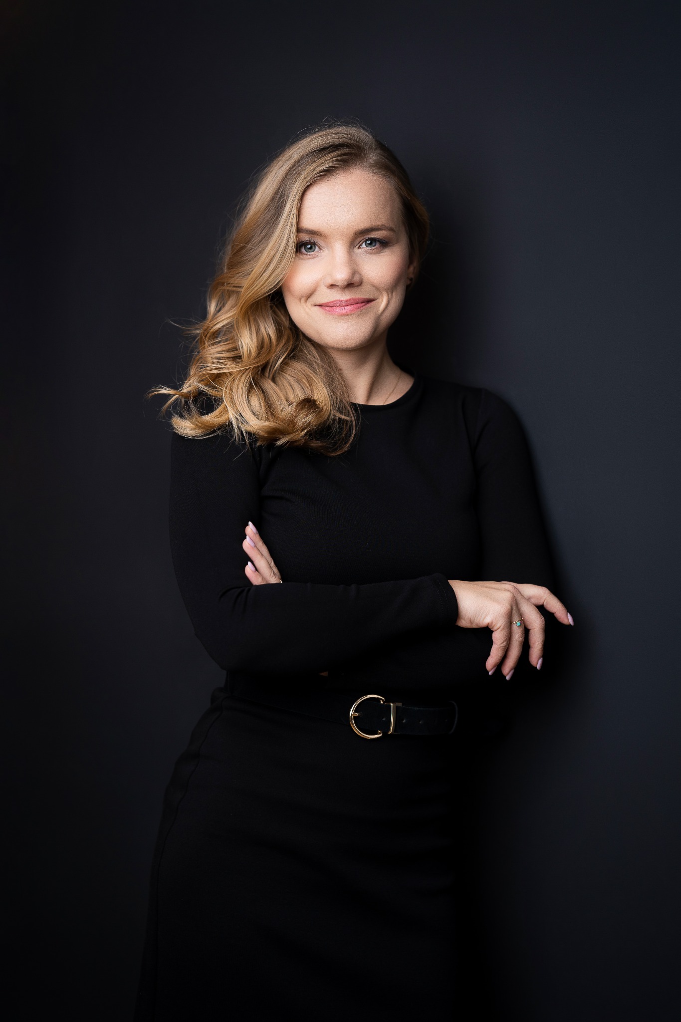 Portret kobiety z blond włosami, ubranej w czarną sukienkę z paskiem z okrągłą klamrą, stojącej na tle ciemnoszarej ściany, z założonymi rękami i subtelnym uśmiechem.