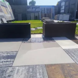 Dwa prostokątne betonowe kwietniki w odcieniach szarości, ustawione na nawierzchni z różnokolorowych płyt chodnikowych, z ekspozycją różnych rodzajów nawierzchni w tle.