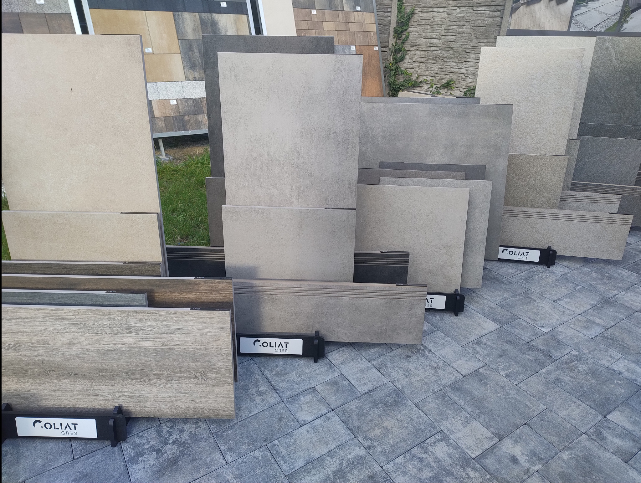 Ekspozycja różnych wzorów i kolorów płytek betonowych na stojakach marki Coliat Gres, ułożonych na nawierzchni z kostki betonowej.