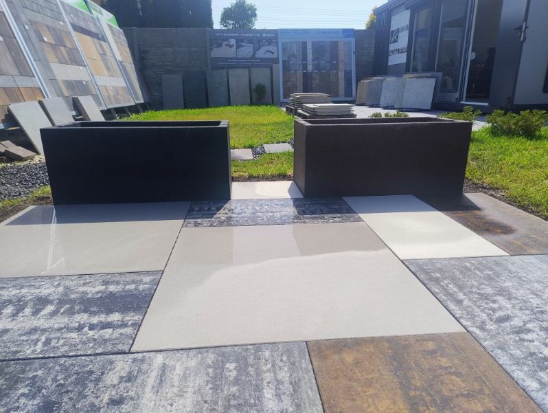 Dwa prostokątne betonowe kwietniki w odcieniach szarości, ustawione na nawierzchni z różnokolorowych płyt chodnikowych, z ekspozycją różnych rodzajów nawierzchni w tle.