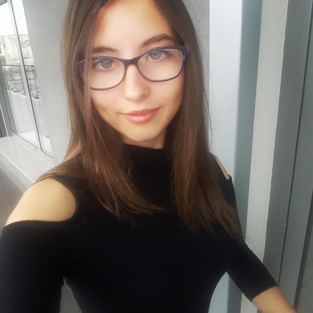 Portret uśmiechniętej młodej kobiety w czarnej bluzce z wycięciami na ramionach i okularach, stojącej na balkonie.