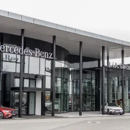 Salon samochodowy Mercedes-Benz z widocznymi modelami aut na zewnątrz, nowoczesna architektura budynku z dużymi przeszkleniami i logo marki.