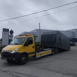 Żółta laweta Iveco z plandeką na platformie, przed salonem Audi w Pruszkowie. Pomoc drogowa z pomarańczowym światłem ostrzegawczym. Transport pojazdu.