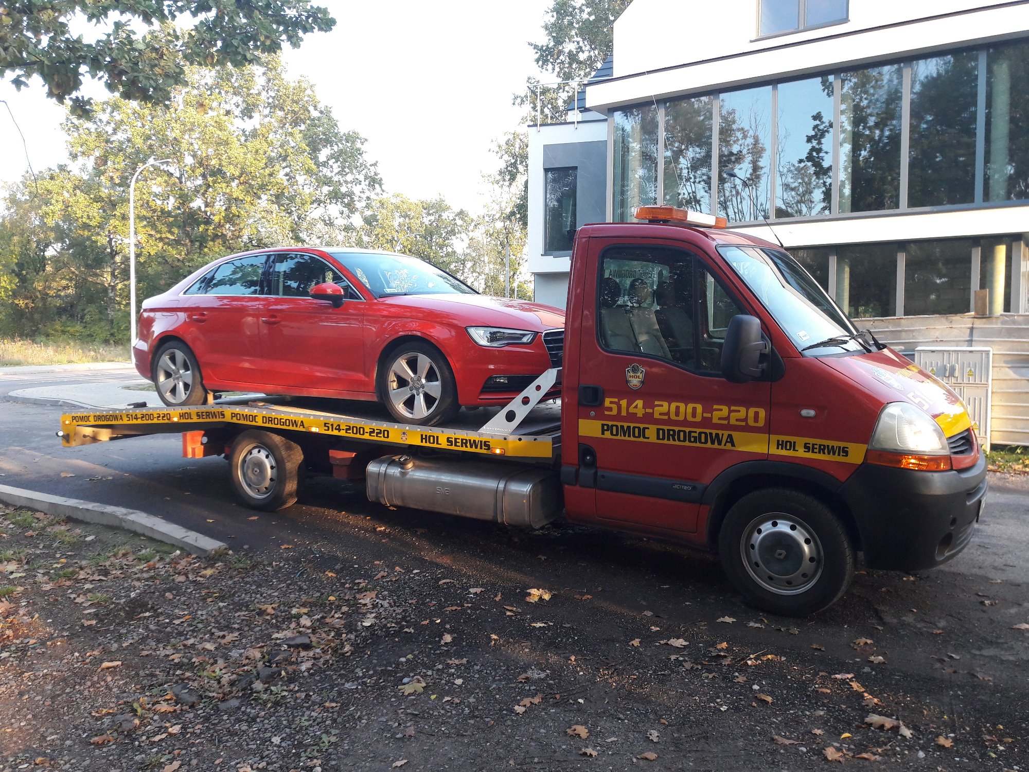 Czerwony samochód osobowy marki Audi przewożony na platformie pojazdu pomocy drogowej z napisem 'POMOC DROGOWA 514-200-220 HOL SERWIS', na tle nowoczesnego budynku.