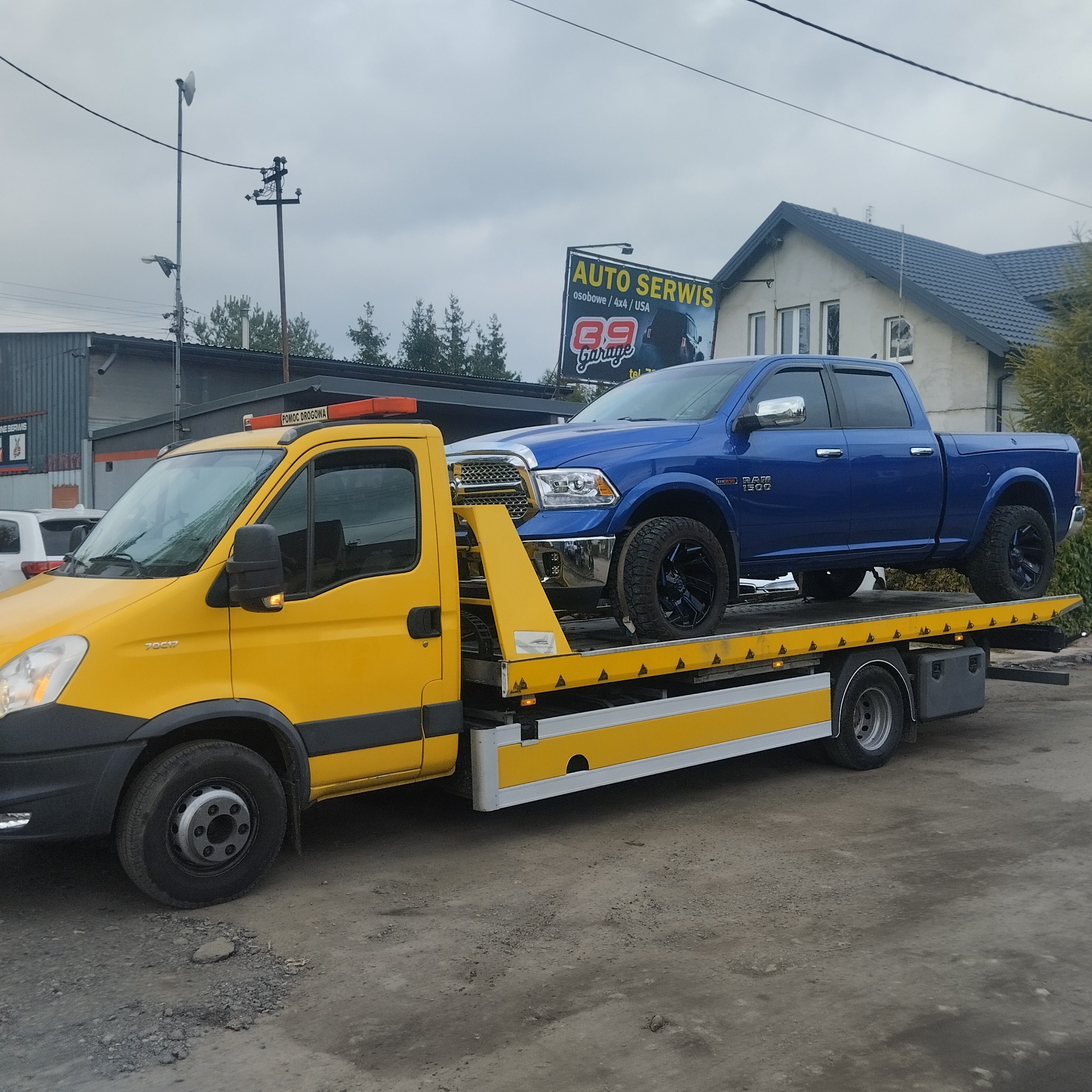 Żółta autolaweta z niebieskim Dodge Ram 1500 na platformie, przed budynkiem auto serwisu. Usługa transportu pojazdów.
