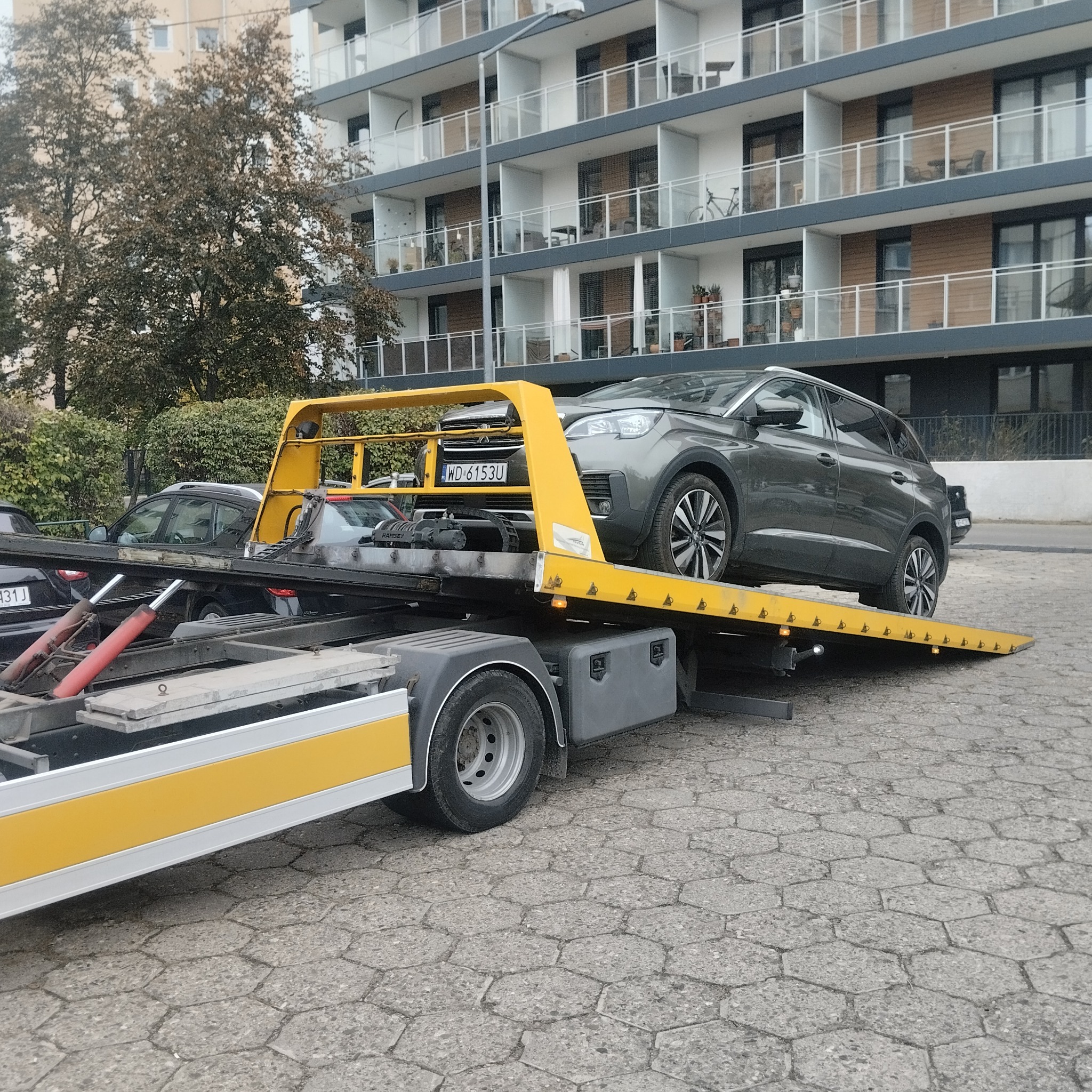 Szary SUV na żółtej lawecie z warszawską rejestracją WD 6153U, na tle bloku mieszkalnego. Usługa lawetowania w mieście.